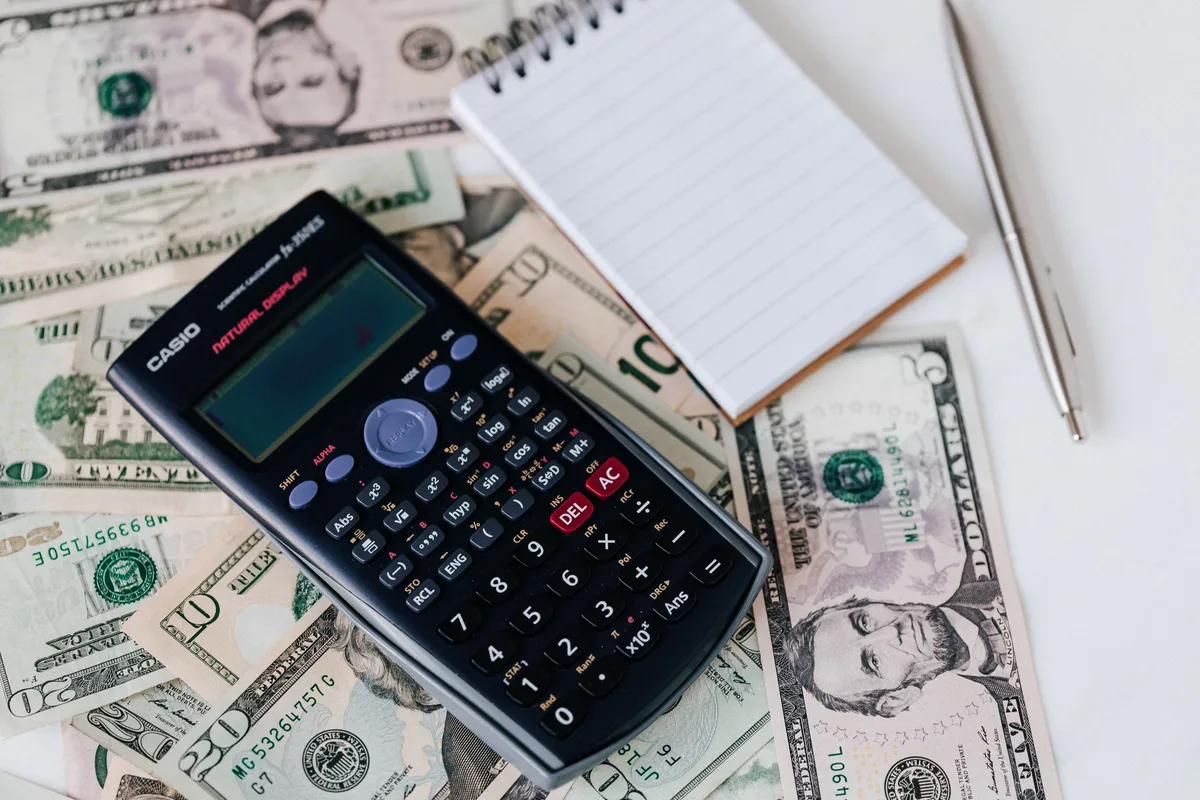 Calculator and notepad placed on stacked dollar bills, representing the running cost of the automation stack