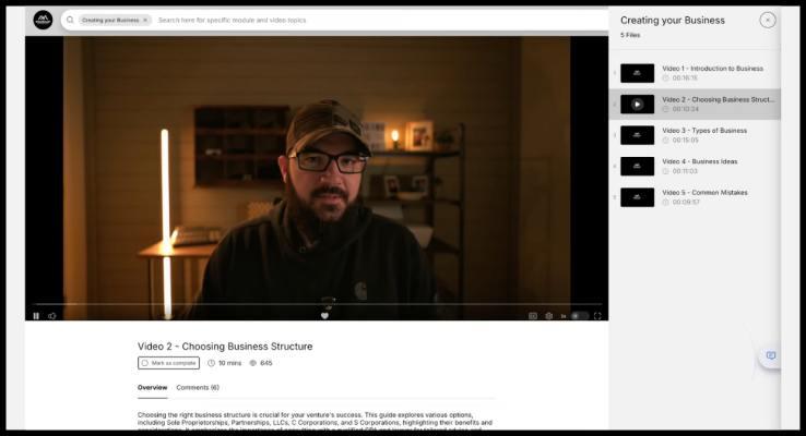 The image shows a video lesson interface titled �Creating Your Business.� On the left is a video player with a presenter in a cozy, dimly lit room. The right panel lists five video lessons, with the second video, �Video 2?�?Choosing Business Structure,� highlighted. Below the video player, there�s an overview and a comments section. The video is 10 minutes long and has 645 views.